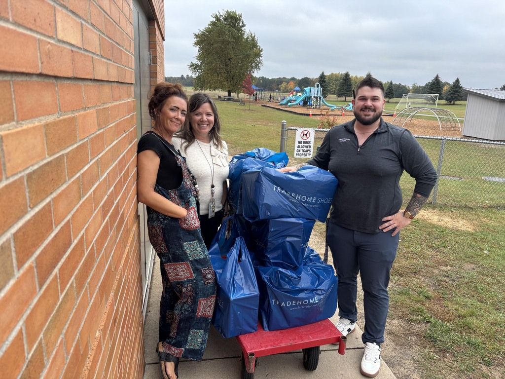 Three people pose next to a cart with blue bags. A building, playground, and grassy field are in the background.