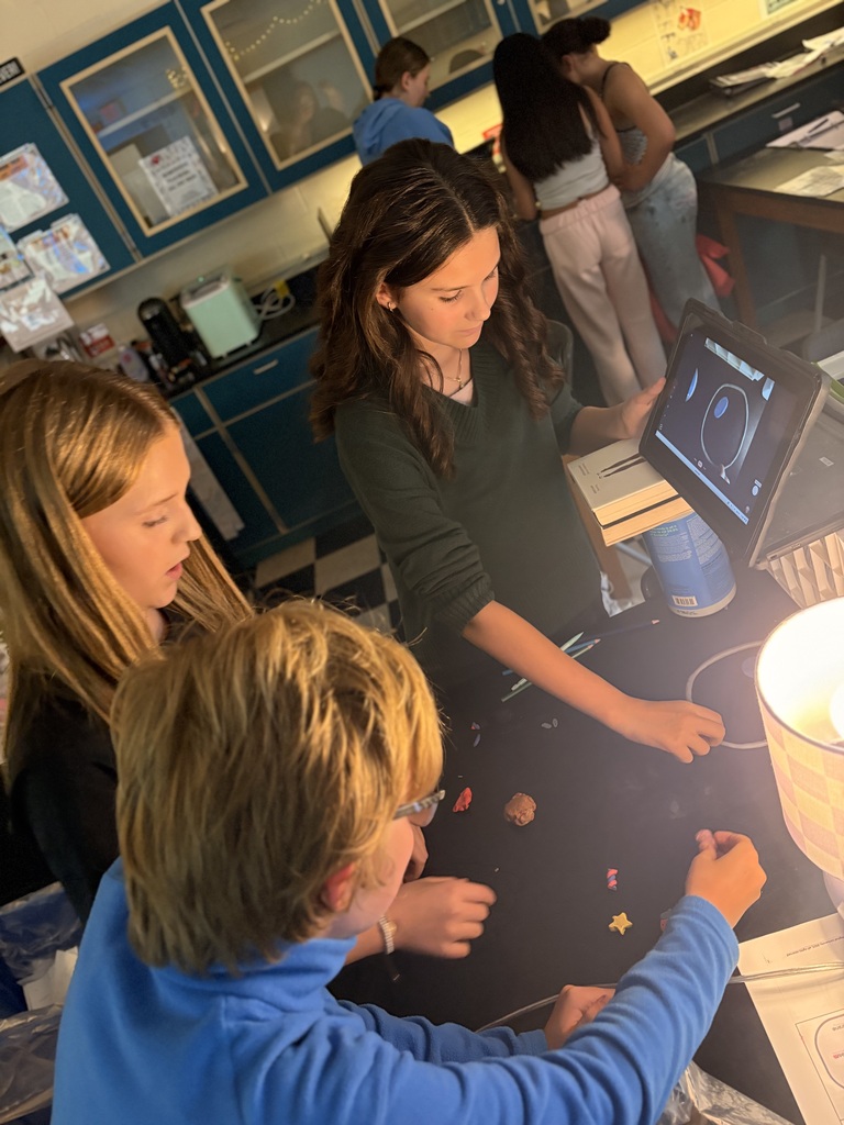 Three students focus on a tablet in a classroom. The girl on the right stands near a desk.