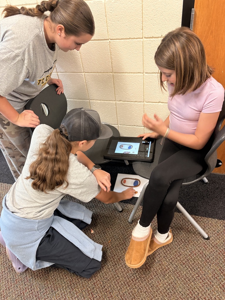 Three girls in a classroom setting. One sitting, one kneeling, and one standing, all interacting with a tablet.