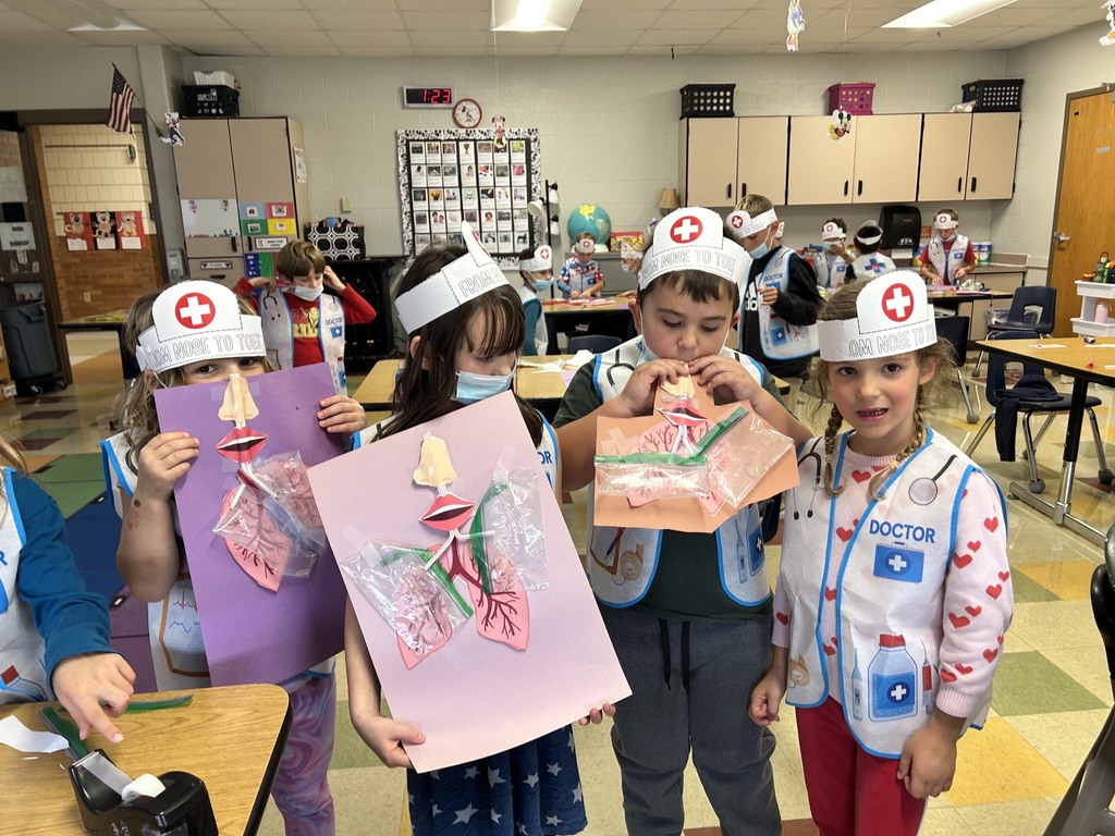 Children in a classroom wearing white hats and masks, holding up paper crafts. A flag is on the wall.