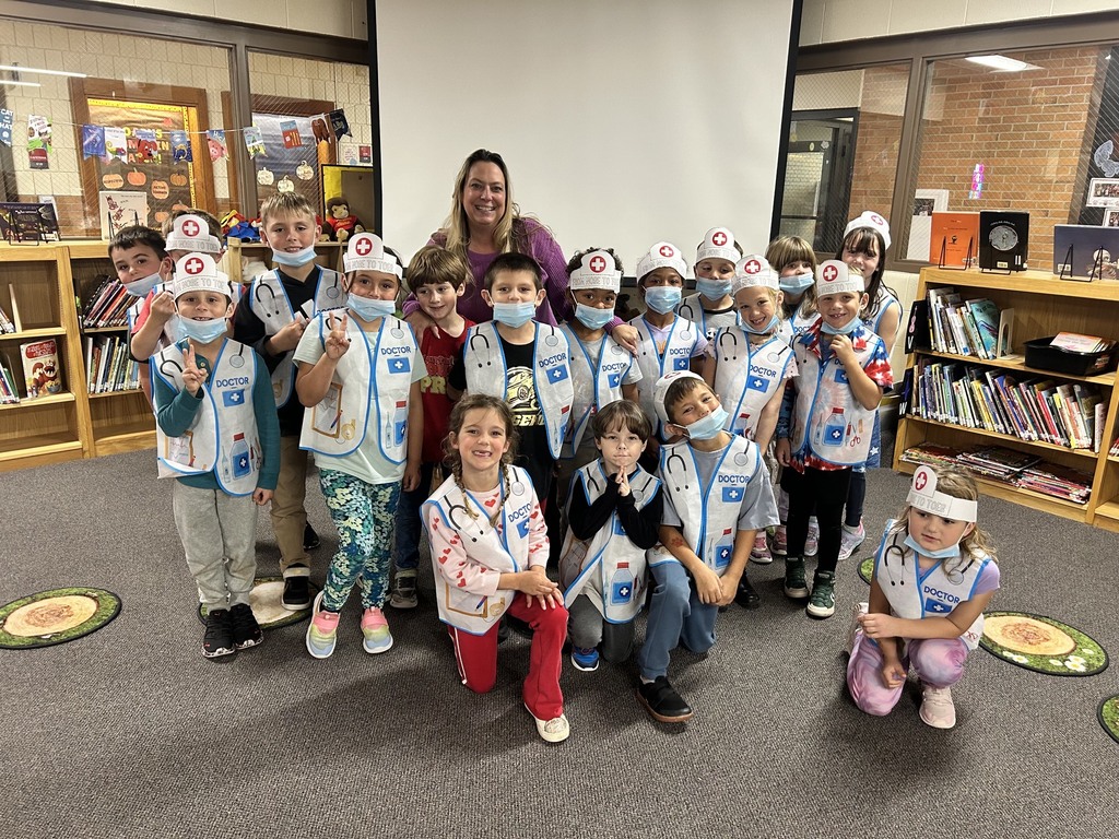 Kids and a woman in a library, all wearing white medical coats, blue hats, and masks.