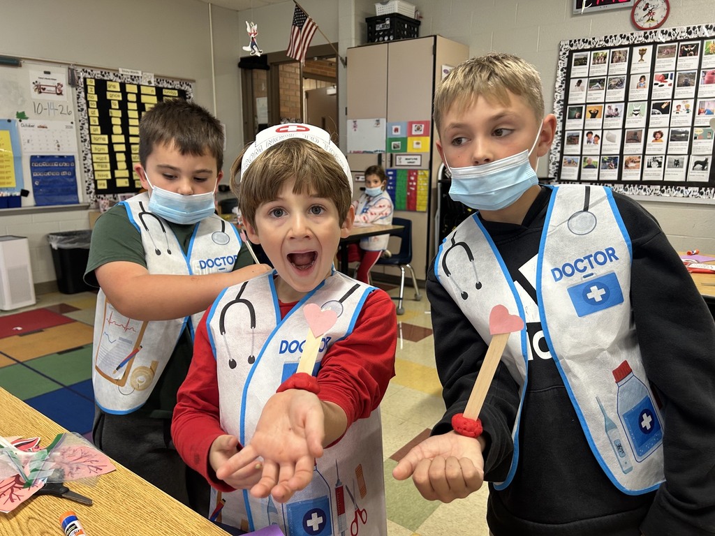 Three children wearing masks and doctor aprons in a classroom. One has a stethoscope around his neck.