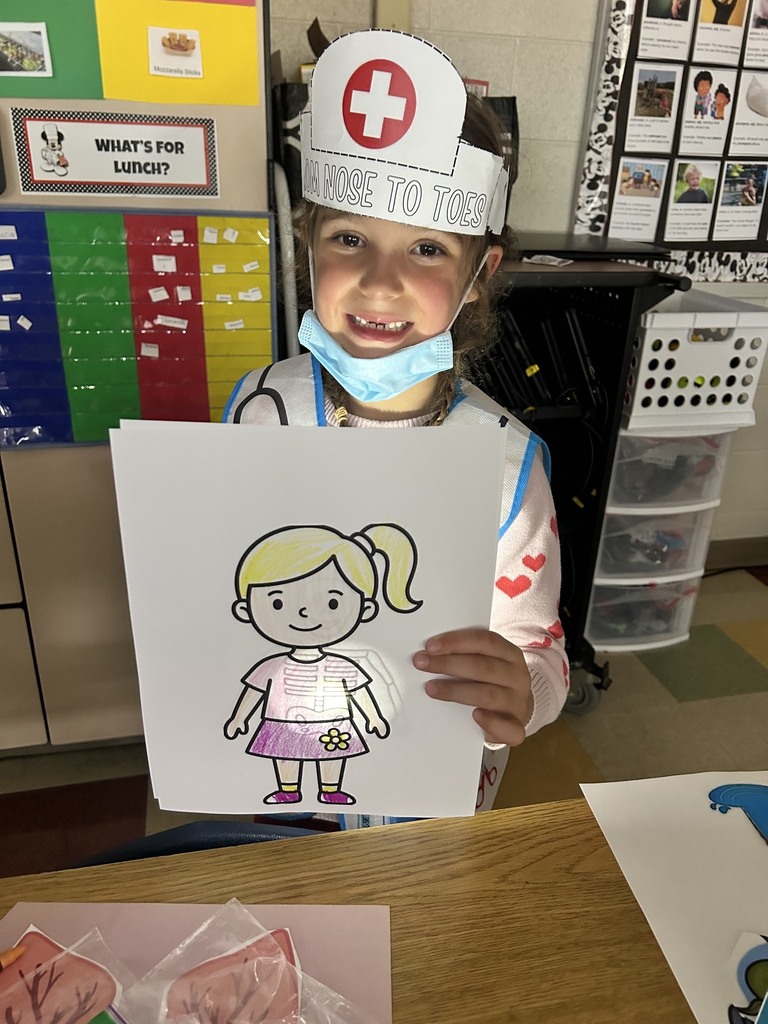 A child in a nurse's hat and mask holds up a drawing of a girl. A classroom background with shelves and storage units.