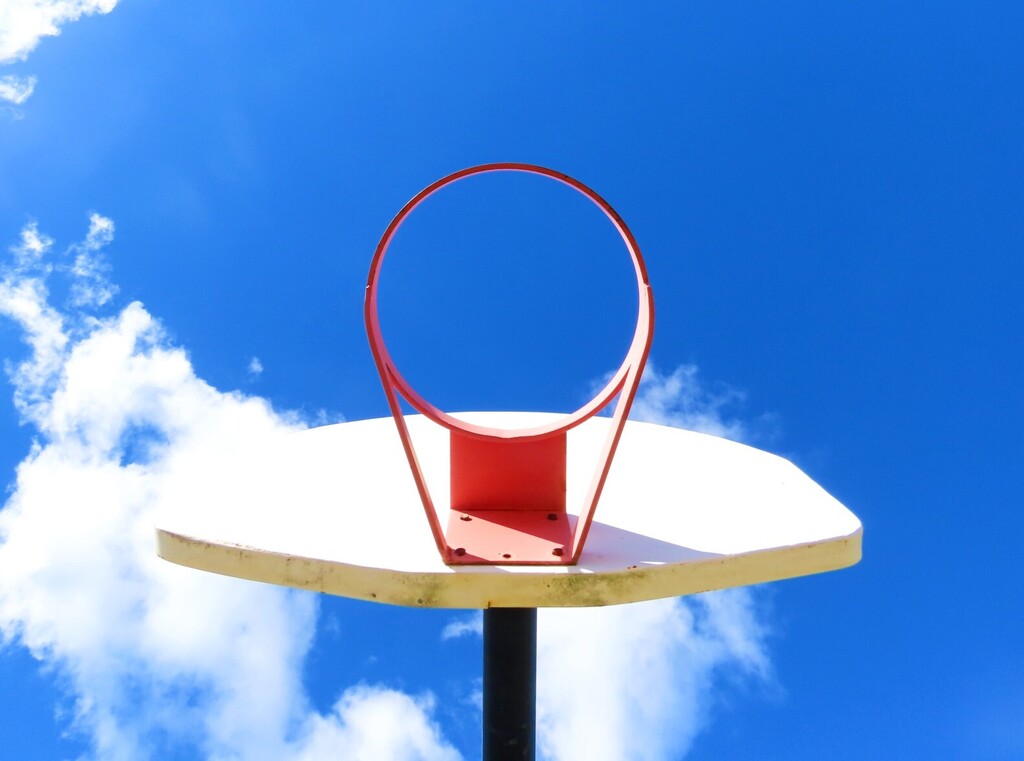 Basketball hoop with red rim and backboard. Bright blue sky with clouds in the background.