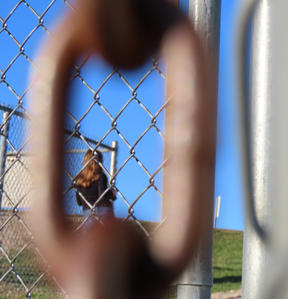 A person with long hair in front of a chain-link fence. Behind the fence, a building is visible. The sky is blue.
