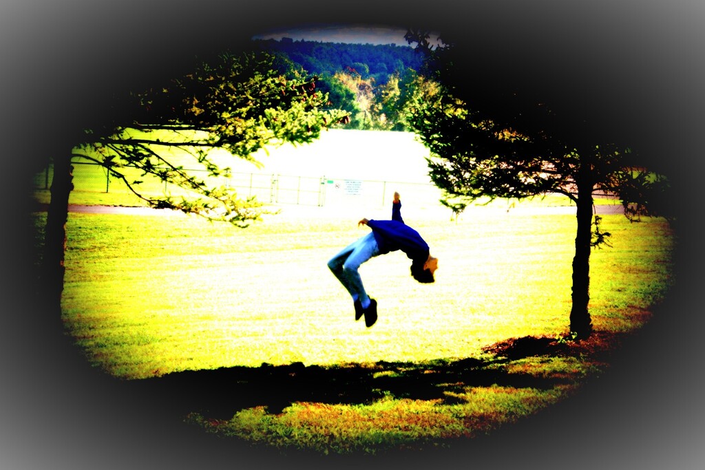 A person is jumping in the air, upside down, in a park with a green lawn and trees.