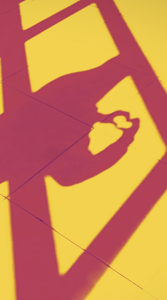 A person's shadow in red on a yellow surface, forming a geometric shape against a tiled floor.
