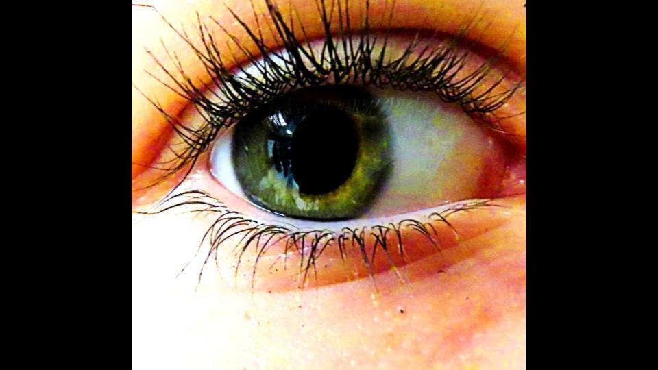 A close-up view of a human eye with long lashes and a green iris, slightly open. The skin around the eye is slightly pink.