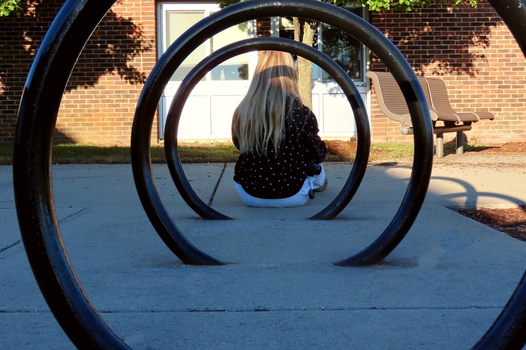 A person with long hair sits on concrete, seen through curved black bike racks. A brick building and a bench are behind.