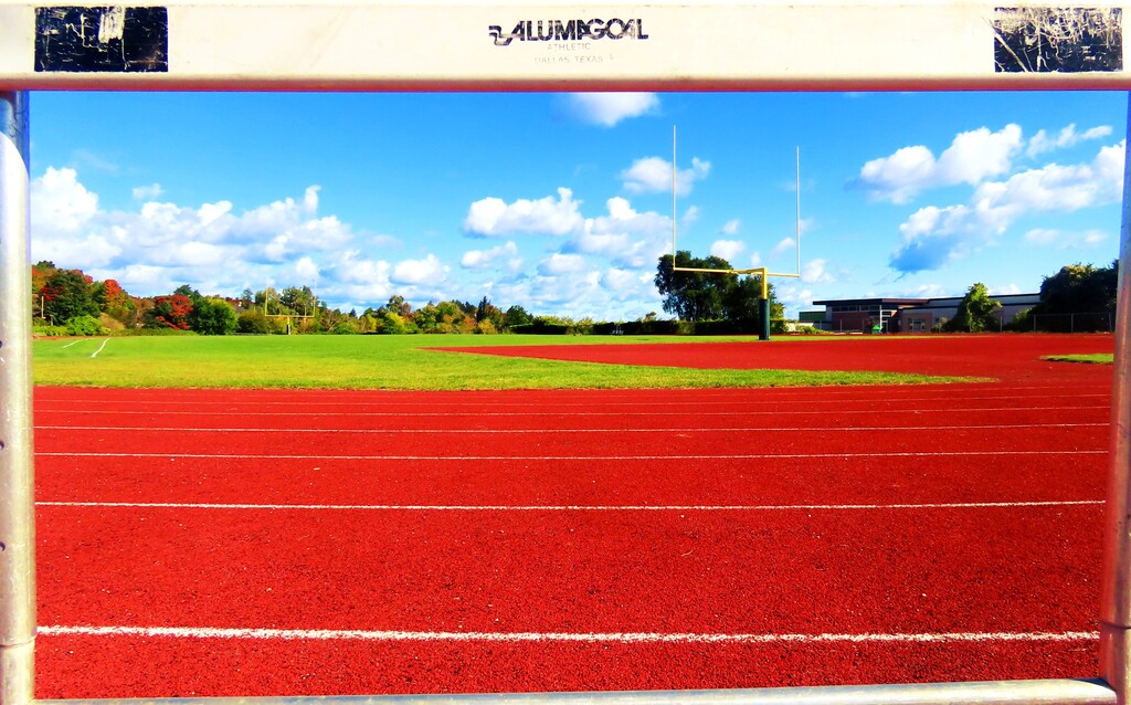 Red track with white lines, green grass, a football field, trees, and blue sky with clouds.