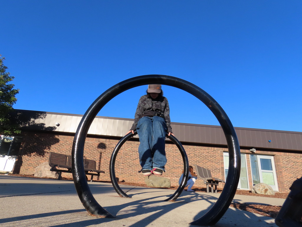 Person in hat and jeans stands on black rings in front of a brick building under a clear blue sky.