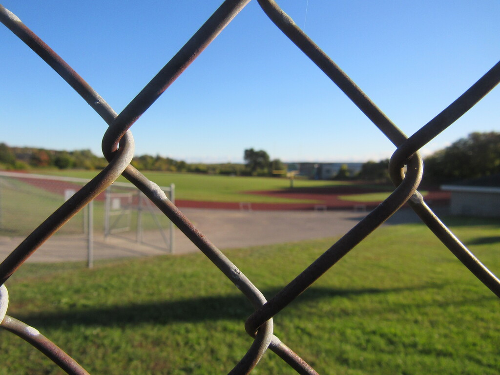 View of a sports field through a rusted chain link fence. The field is empty.