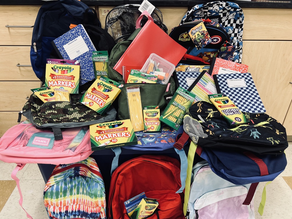 Multiple backpacks with colorful designs, some open and filled with school supplies, against a backdrop of wooden cabinets.