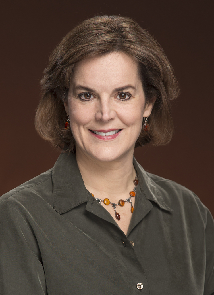 Woman with short brown hair, smiling. She wears a gray shirt, a necklace with orange and brown beads, and earrings. Brown background.