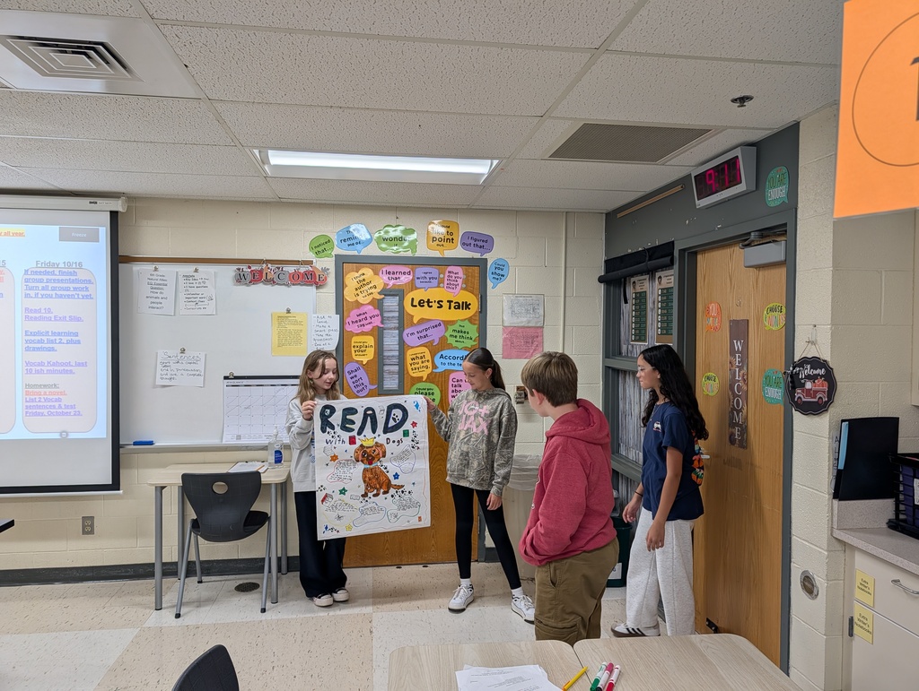 Five students in a classroom. One holds a large sign that says 'READ'. Behind them, a projector screen is active.