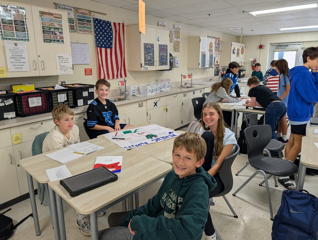 Students work at tables in a classroom. An American flag is in the background. A few students stand in the background.