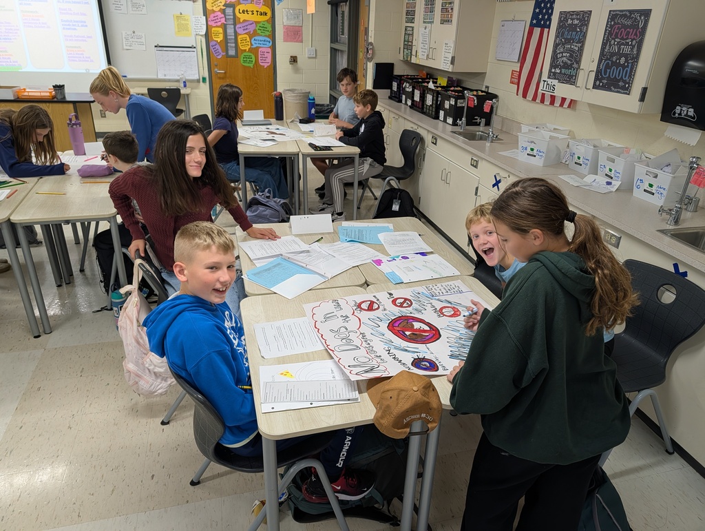 A classroom with students seated at tables. One group of students is gathered around a table with papers.