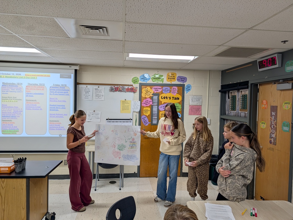 Four students in a classroom, one standing and holding a whiteboard, three others are listening.