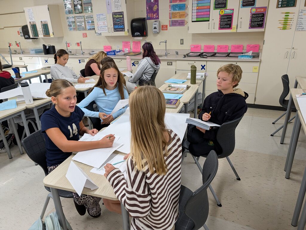 Several children in a classroom, seated at tables, discussing and examining papers. White cabinets line the walls.
