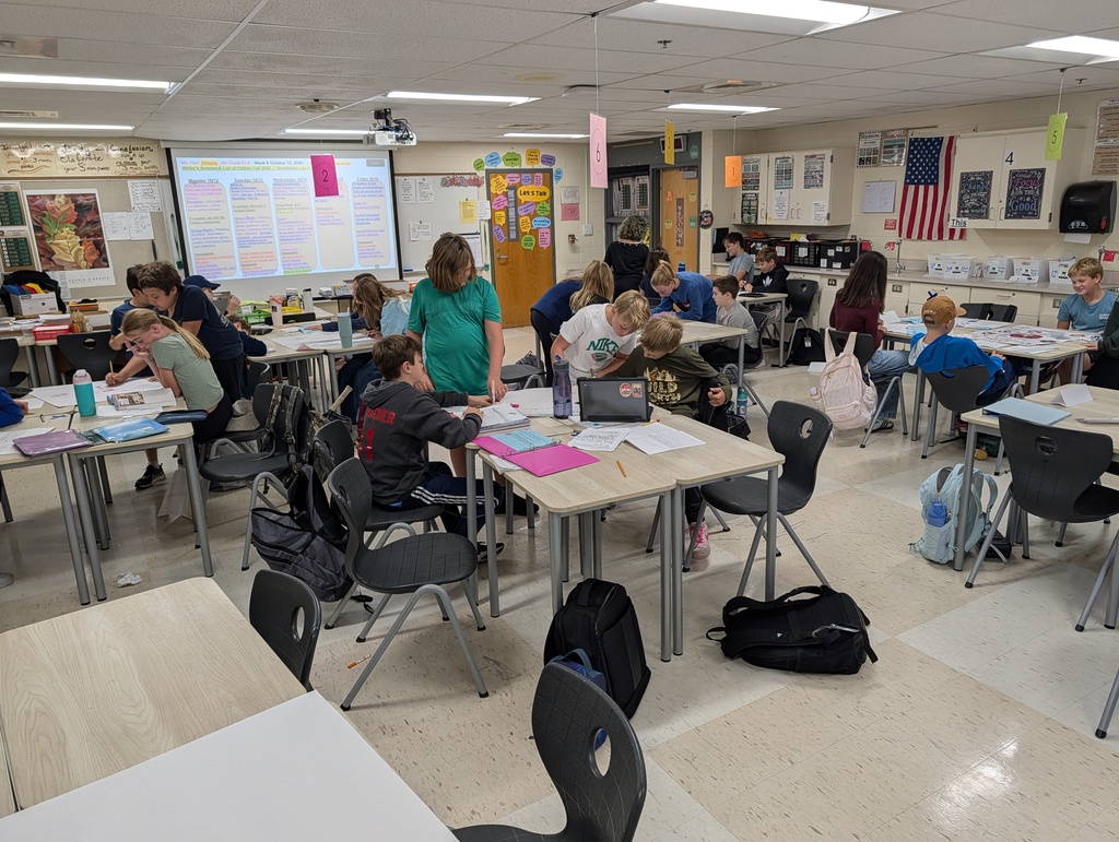 A classroom with tables and chairs, where students are sitting and working on papers. A teacher is standing at a desk, and a projector screen is visible.
