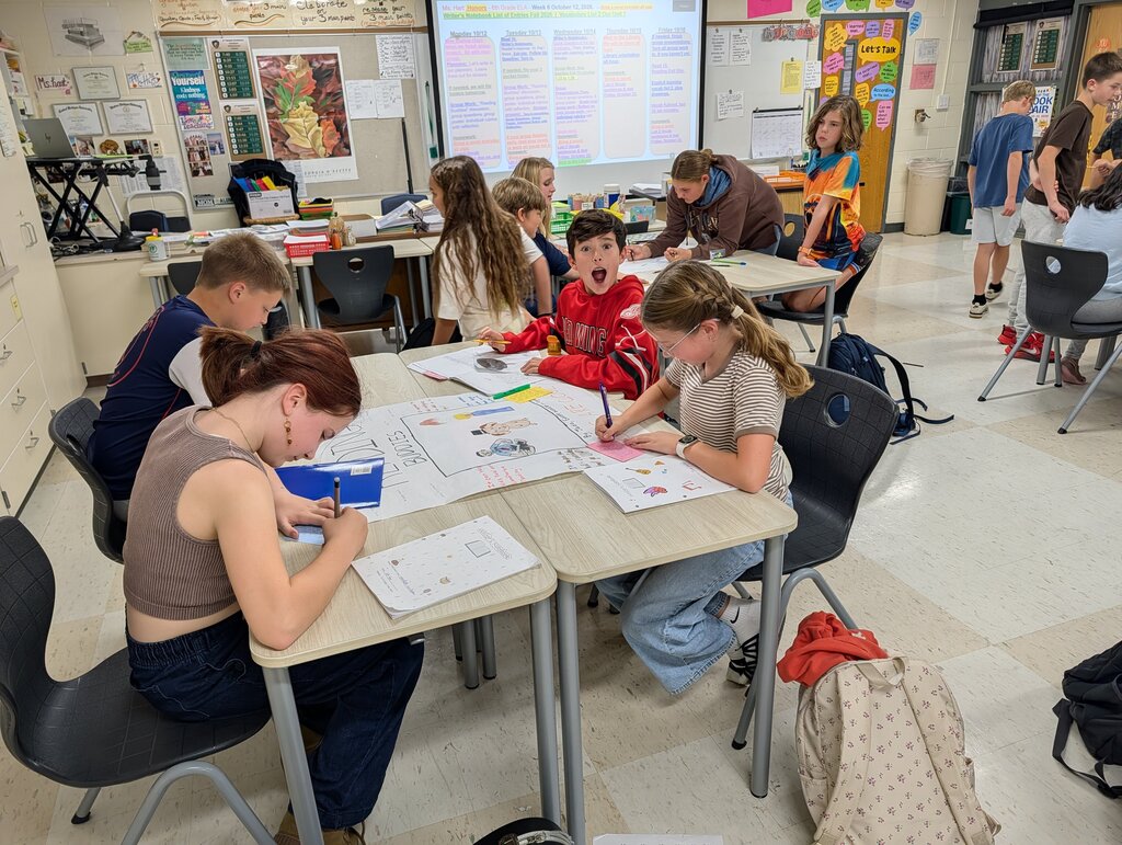 Group of students in a classroom. One student stands behind a table with a projector screen on the wall.