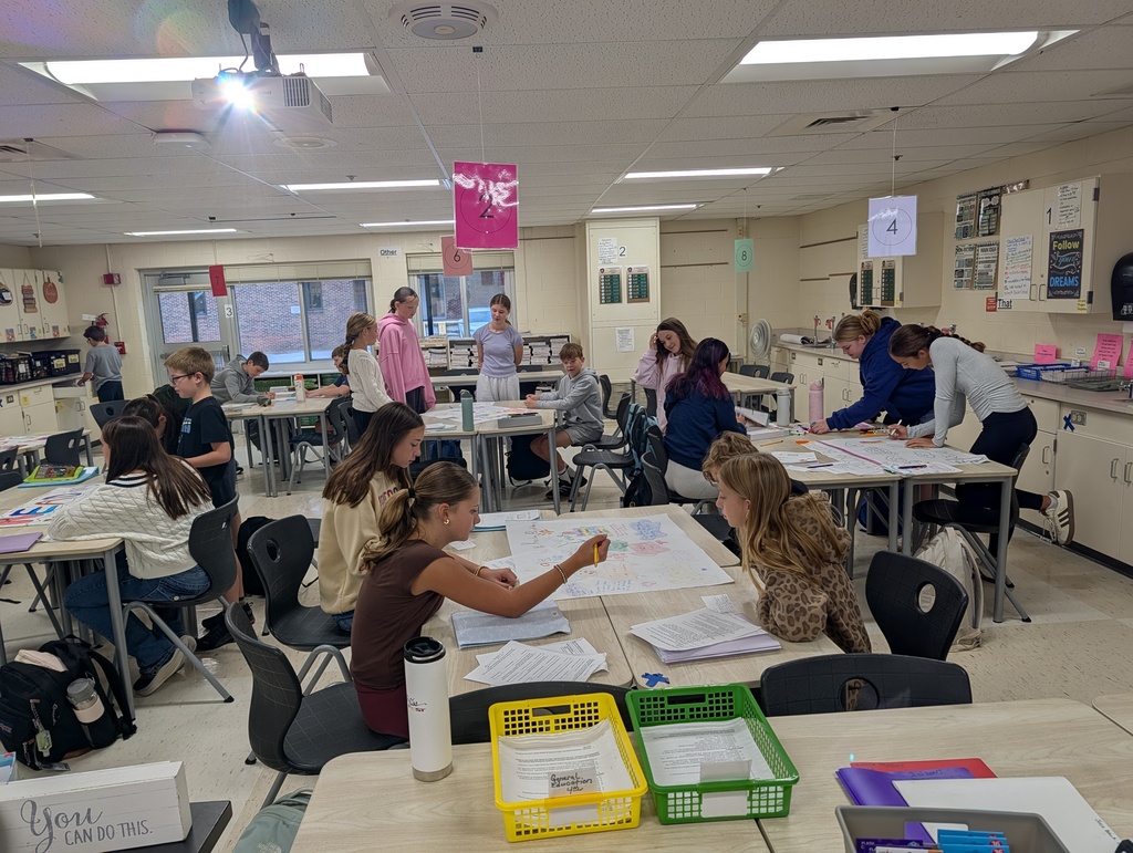 A classroom with tables where students sit at desks and work on projects, and fluorescent lights on the ceiling.