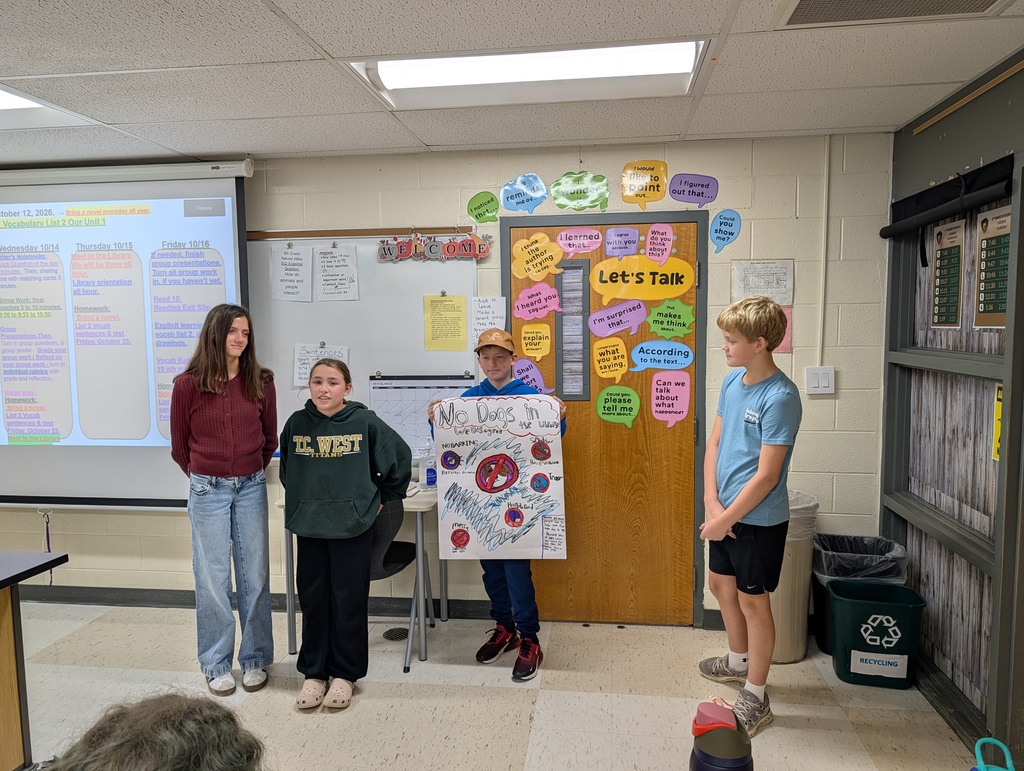 Four children, two girls and two boys, stand in a classroom with a projector screen. One child holds a poster.