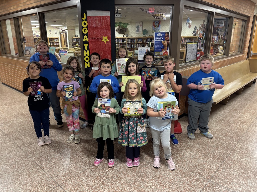 Group of children in a hallway with books, smiling. Behind them, a library with shelves and a bench.