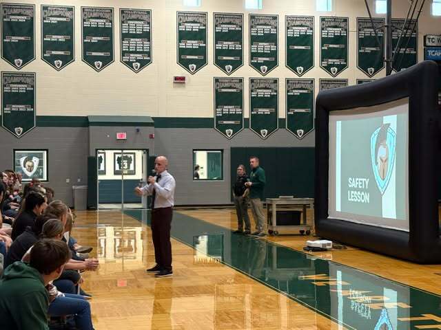A man in a white shirt stands in a gym, speaking into a microphone in front of a projector screen.
