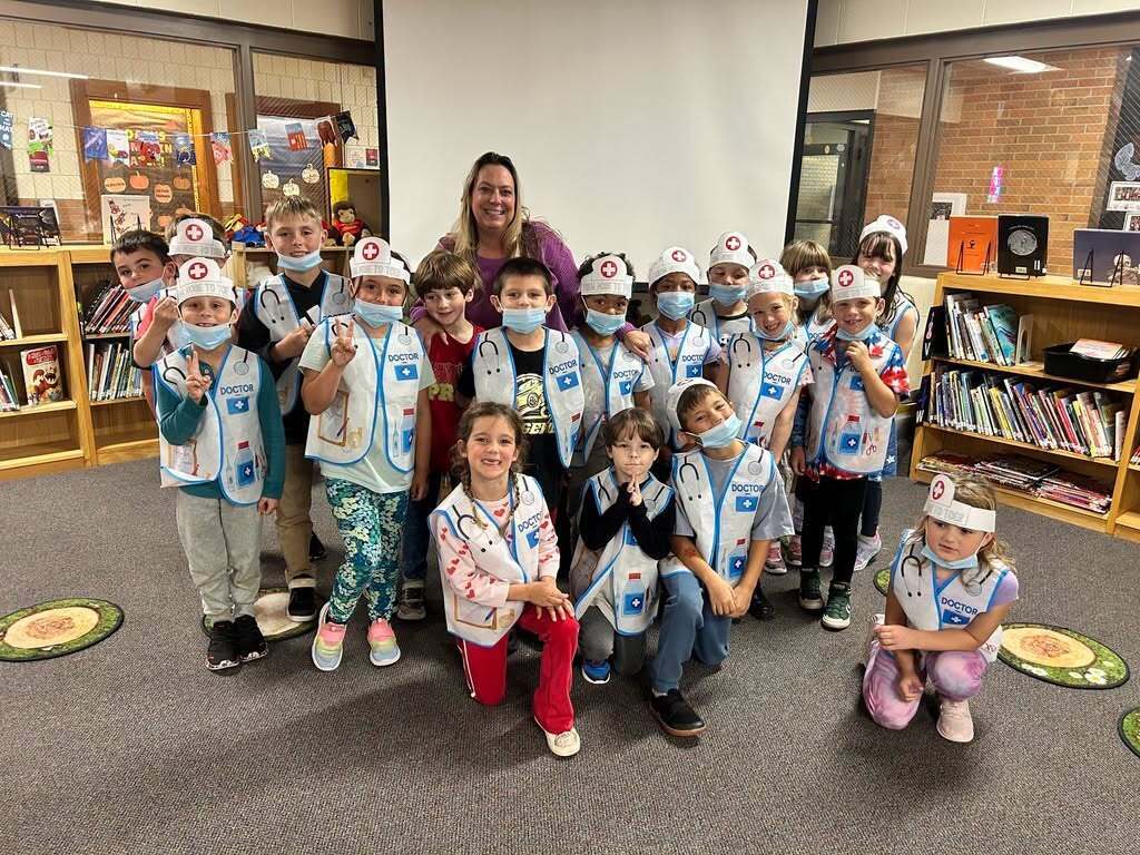 A group of young children wearing blue vests and masks pose for a photo in a classroom.