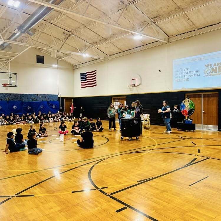 An indoor basketball court with children seated, two adults standing at the front, and a large projector screen on the wall.