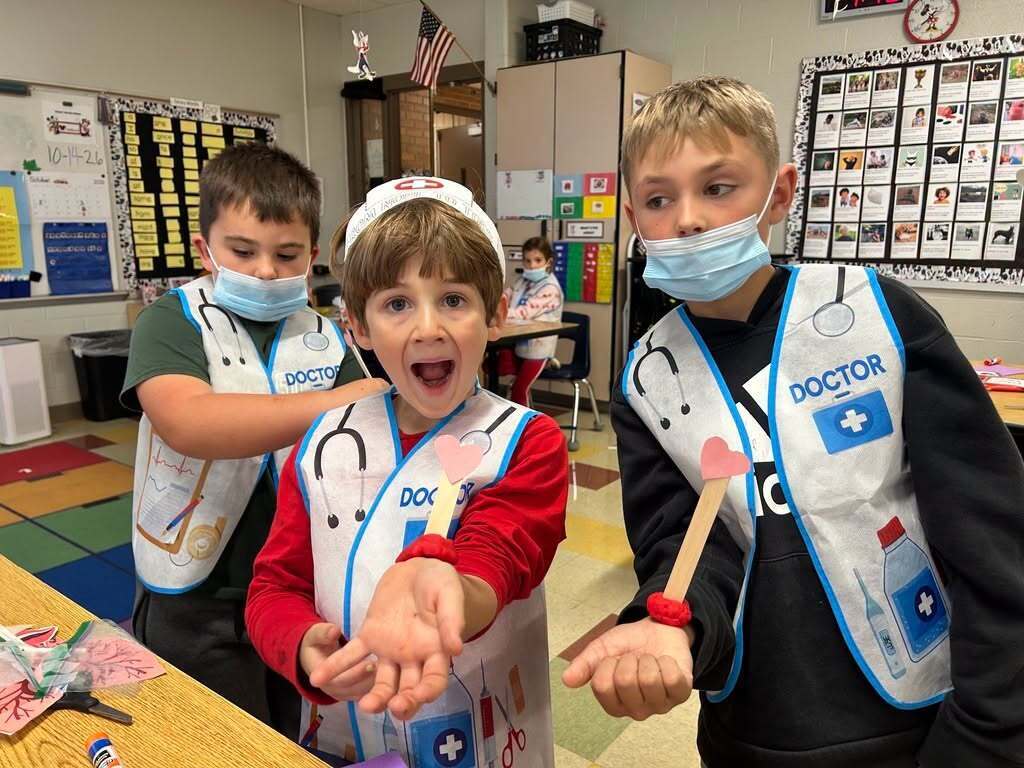 Three boys in a classroom wear lab coats and masks; one holds a toy drum and smiles.