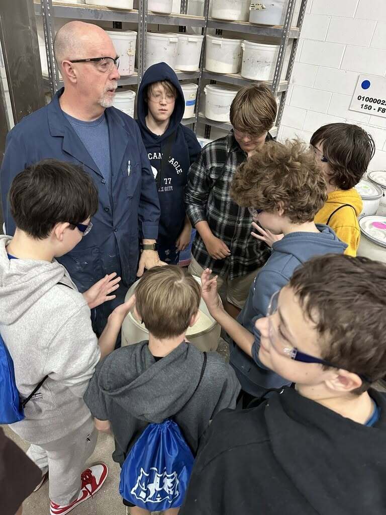 An adult man in a blue jacket is surrounded by students; they are looking at a large metal pot.