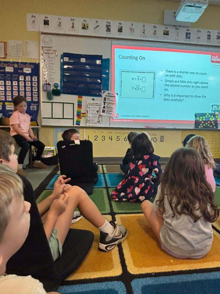 Classroom with projector screen displaying math problem. Children sit on floor or chairs, facing screen, listening to teacher.