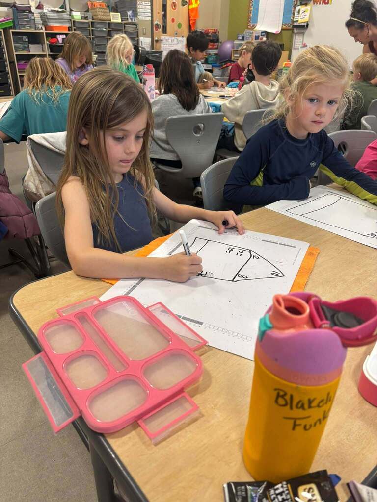 Two young girls drawing on a piece of paper at a table in a classroom.