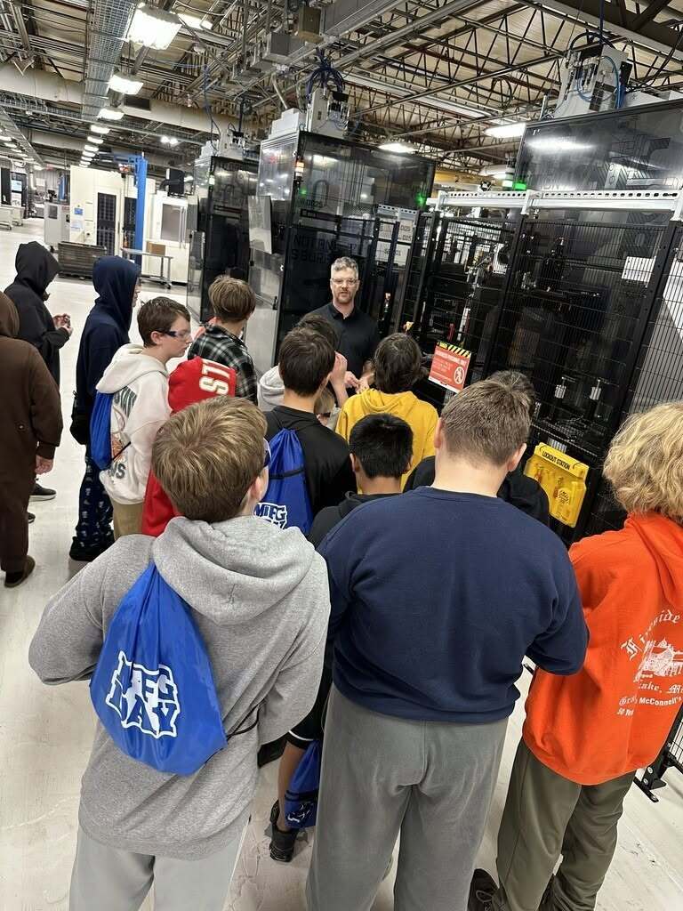 Students observe a presentation about machines inside a factory with large glass doors.