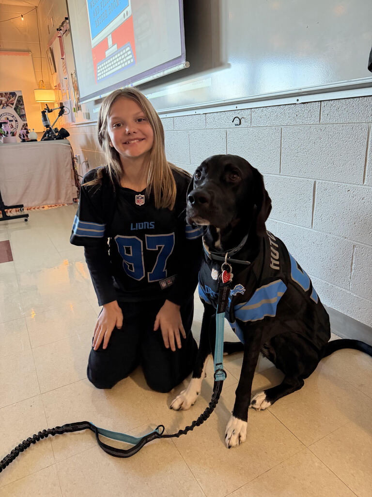 Girl wearing a Detroit Lions jersey kneeling beside a black dog in a blue vest with white paw prints.