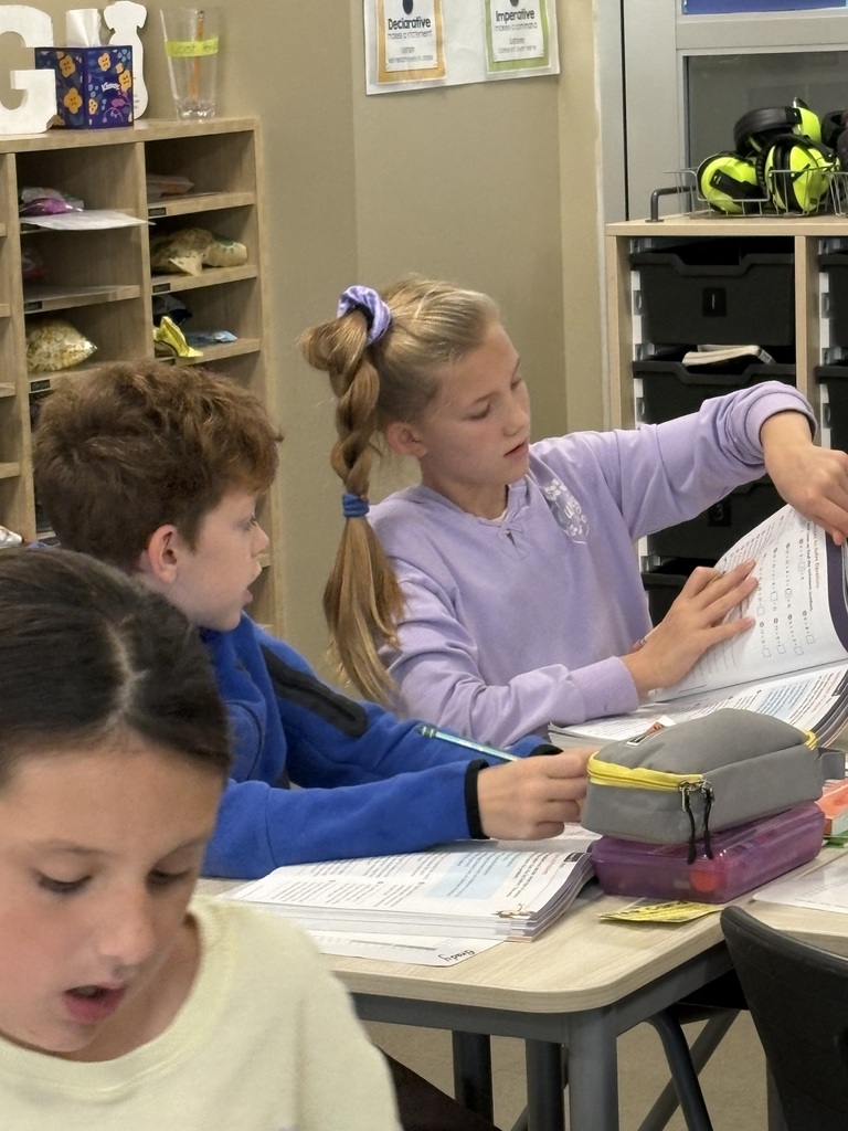 Students seated at desks in a classroom, looking at math books and papers; shelves with books in background.