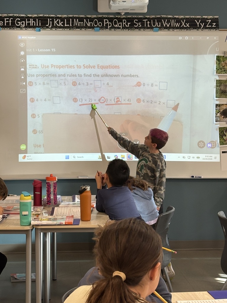 Children seated in classroom, one standing with pointer, pointing at screen during a math lesson. Screen shows alphabets and arrows.