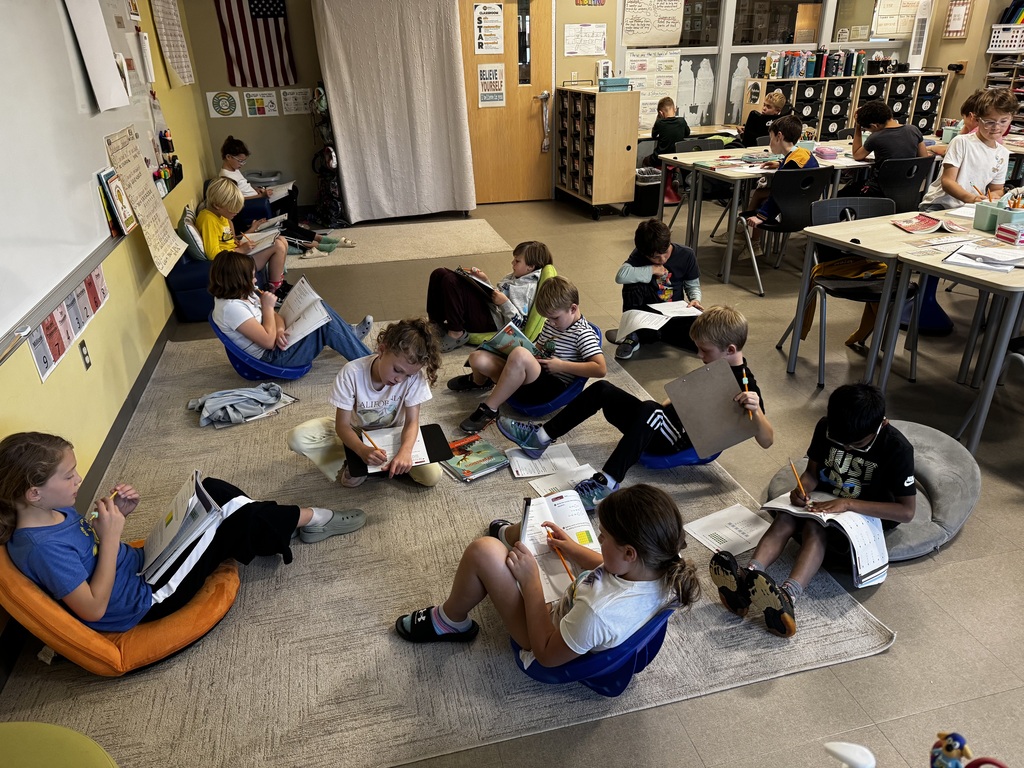 Students in a classroom, sitting on the floor with bean bags and books, working on math.