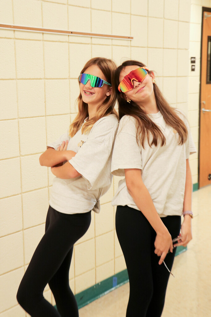 Two girls in white T-shirts and black leggings stand in front of a tiled wall. Both wear large multi-colored sunglasses.