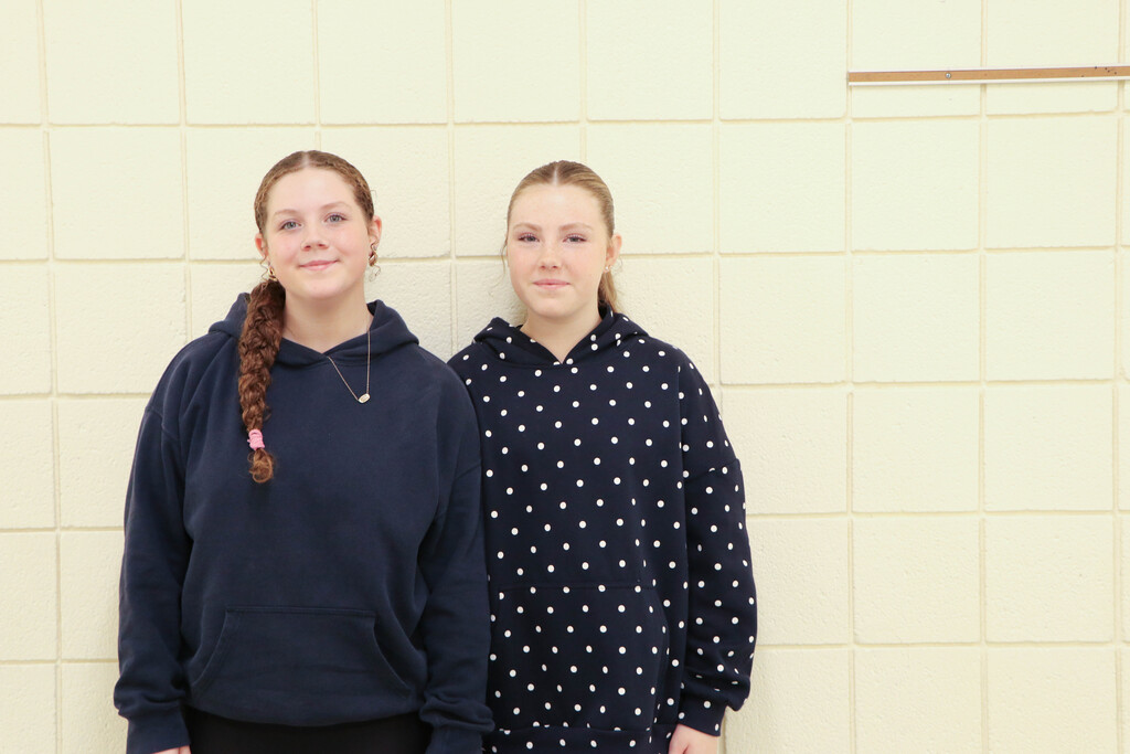 Two girls stand against a yellow tiled wall, one in a navy hoodie with a braided ponytail, the other in a polka-dot hoodie.