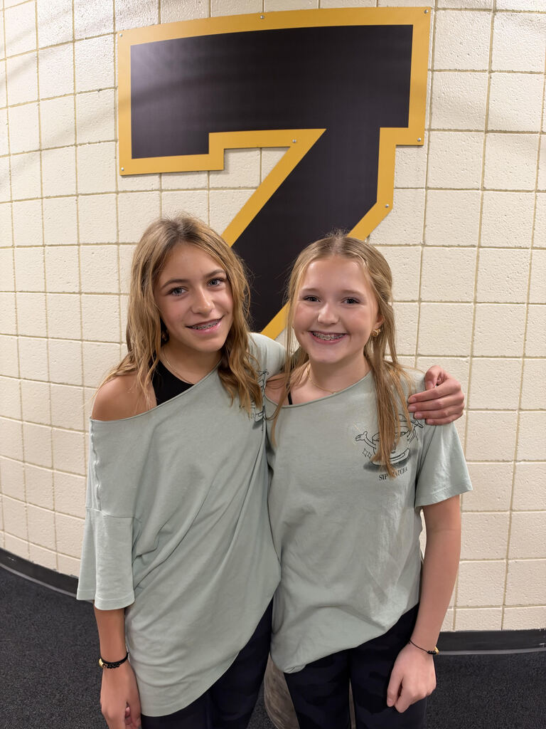 Two girls in matching gray shirts, standing close with arms around each other, pose in front of a white wall.