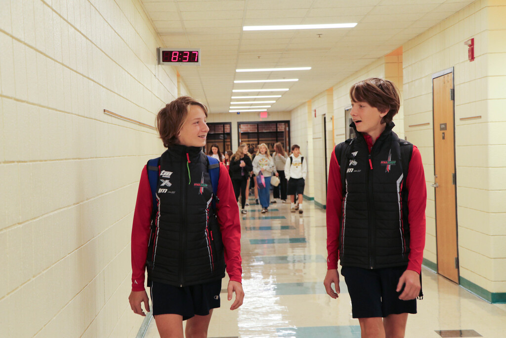 Two students in athletic vests walk in a hallway; a clock and doors are on the wall.