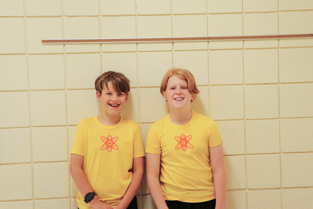 Two young boys wearing yellow shirts with red atom designs, standing next to each other against a tiled wall.