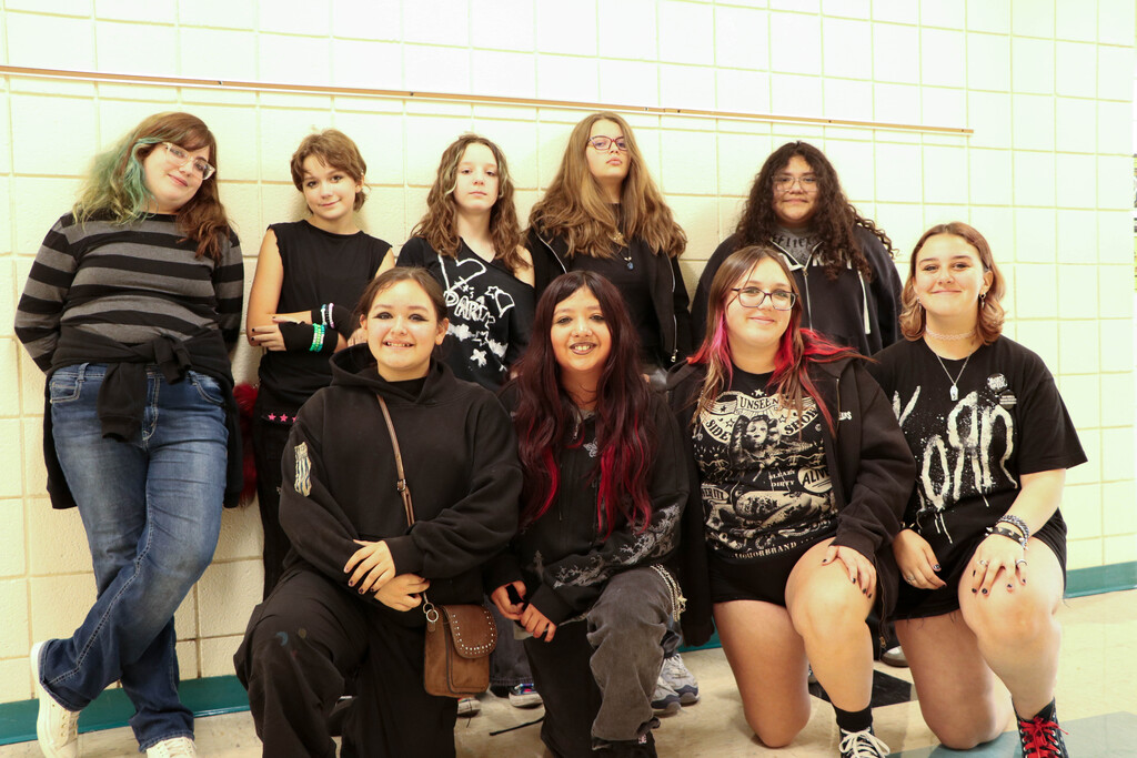 A group of young people smile, with one crouching, against a white tiled wall.