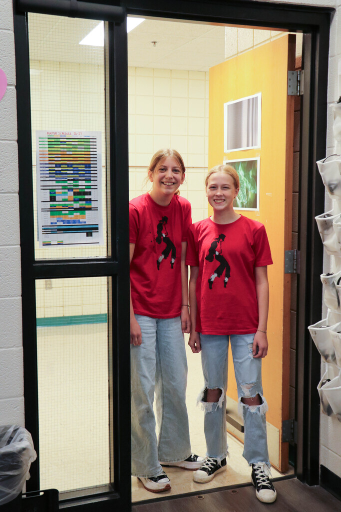 Two girls in matching shirts pose by a door with a yellow wall and a bulletin board.