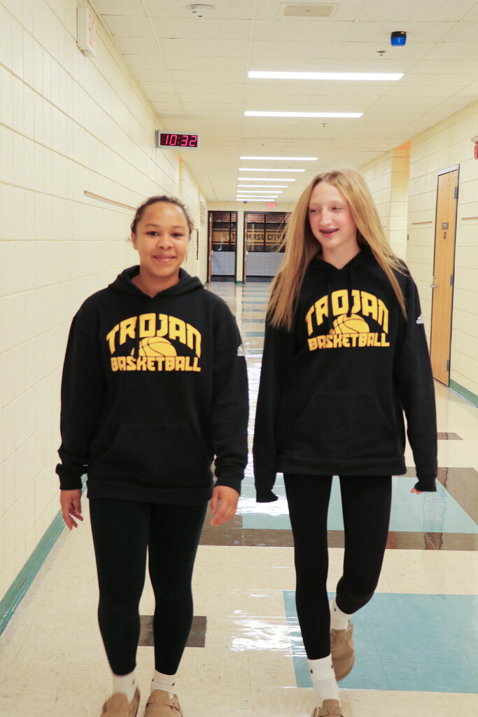 Two students stand in a school hallway. They wear black hoodies and leggings. Both have shoes.