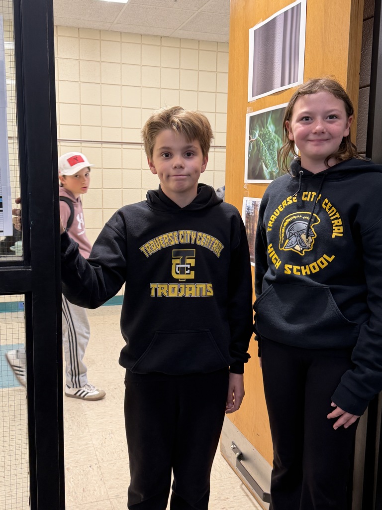 Two children in school sweatshirts stand at a door with another child and pictures on a wall.
