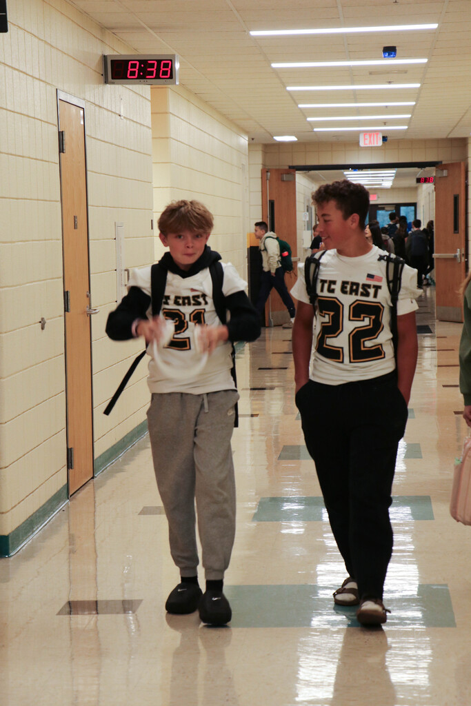 Two students in athletic jerseys walk down a school hallway. One wears black shoes and the other wears white shoes.